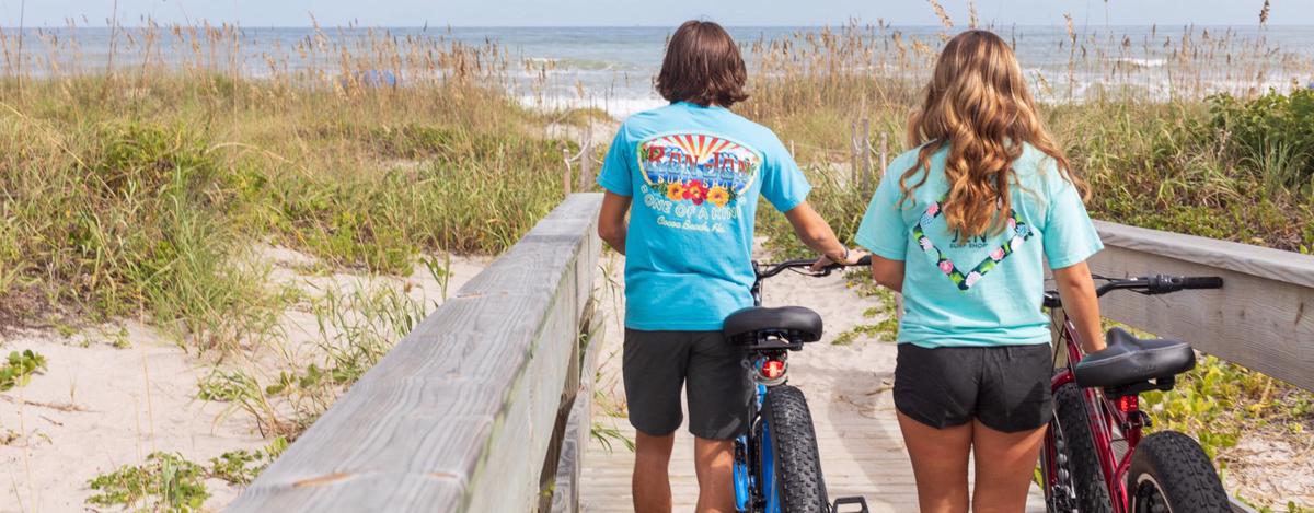 two people wearing short sleeve t-shirts walking bicycles on a boardwalk