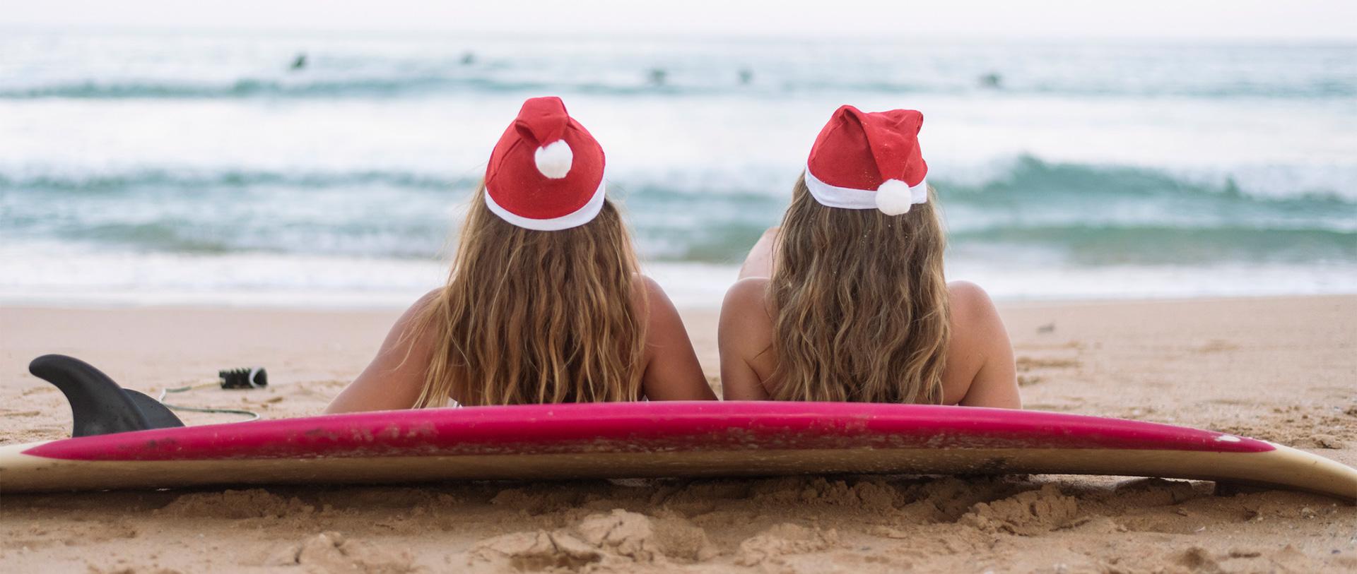 two girls relaxing against a surfboard while wearing red santa hats