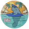 Cartoon manatee with sunglasses and blue surfboard in water, surrounded by palm trees, beach, and sunset. Ron Jon Surf Shop logo at the bottom left.