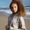 A woman with curly hair stands on a beach wearing a light-colored camo hoodie, looking to the side with the ocean and sand in the background.
