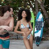 Two people in swimwear smiling near surfboards by a tree-lined fence.