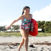 Young girl in a floral swimsuit holding a red bodyboard walks on a sandy beach with vegetation in the background.