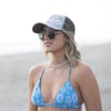 A woman wearing a blue floral bikini top, sunglasses, and a patterned cap stands on a beach.