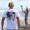 A person wearing a white t-shirt with a tropical graphic stands on a beach, while another person in the background plays paddleball near the shore.