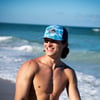 Shirtless young man wearing a blue patterned baseball cap stands on a beach, smiling, with the ocean and sky in the background.
