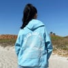 A person with a high ponytail wearing a light blue hoodie with a sun and wave graphic stands on a sandy beach facing away from the camera.