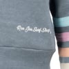 Close-up of the Ron Jon Surf Shop logo in white, against a gray-blue background. Stripes in teal, purple, dark gray, and pink are visible on the arm.