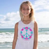 A young person stands on a beach wearing a white sleeveless shirt with a colorful peace sign and flower design, smiling at the camera. Waves and blue sky are in the background.