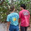Two people walk on a forest path. The woman wears a mint Ron Jon Surf Shop t-shirt, and the man wears a pink Ron Jon Surf Shop t-shirt. Trees and foliage surround them.