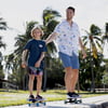 An adult and a child ride skateboards on a sidewalk in a sunny park with palm trees in the background.