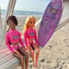 A doll stands on the beach next to a purple surfboard, a toy turtle, a pink vest, sandals, and miniature beach accessories with the ocean in the background.