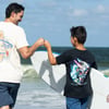 Two people holding a surfboard near the ocean fist bump each other, wearing casual t-shirts and shorts.