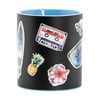 A black mug with a light blue interior, decorated with colorful stickers including a surfboard, cassette tape, turtle, pineapple, hibiscus flower, and a surf-related sign.