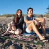 Two people sit on a blanket at the beach, smiling. The blanket has “Ron Jon Life Shop” written on it. Grass and blue sky are visible in the background.