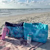 Two patterned beach bags and two tumblers sit on a towel on the sand, with the ocean and a partly cloudy sky in the background.