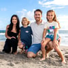 A family of four poses and smiles together on a sandy beach with the ocean and blue sky in the background.