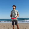 A man stands on a sandy beach holding a red frisbee, wearing a light gray T-shirt and dark shorts, with the ocean and blue sky in the background.