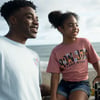 Two people smiling near a beach. The person on the left is wearing a light blue shirt, and the person on the right in a pink Surf Shop shirt is sitting on a railing.