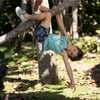 Child hanging upside down from a tree branch, wearing a turquoise tank top, patterned shorts, and checkerboard shoes, with trees and foliage in the background.