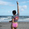 Person on a beach wearing a gray shirt with a colorful design and pink swim bottoms, facing the ocean and raising one arm with a peace sign.