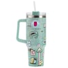 A large mint-green travel tumbler with a handle, straw, and whimsical illustrations of a frog, snail, mushrooms, flowers, and a pink box character on the exterior.