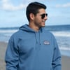 A man wearing sunglasses and a blue hoodie stands on a sandy beach with the ocean and cloudy sky in the background.