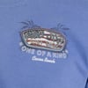 A blue shirt with a Ron Jon Surf Shop One of a Kind Cocoa Beach logo featuring an American flag background on the chest.