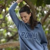 A woman wearing a blue RON JON sweatshirt stands outdoors, smiling and leaning her arm on a tree branch, with green leaves in the background.