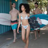 Three people in swimwear carry surfboards while walking along a boardwalk near a beach.
