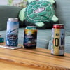 Three canned drinks in colorful Ron Jon Surf Shop can coolers are placed on a wooden table, with a turtle-shaped Ron Jon pillow on a couch in the background.