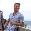 Man in floral shirt leans on a wooden railing, smiling and looking off to the side. Ocean and foliage are in the background under a cloudy sky.