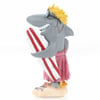 A cartoon shark figurine wearing sunglasses, yellow hair, a pink grass skirt, and holding a red and white surfboard, standing on a sandy base.