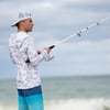 A person wearing a white camo hoodie, blue shorts, and a backward cap is fishing by the ocean on a cloudy day.