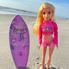 A blonde doll in a pink crop top and shorts stands on sand beside a pink surfboard with floral designs and Ron Jon Surf Shop logo. The beach and ocean are in the background.