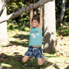 Child in a blue shark tank top and patterned shorts hangs from a tree branch, smiling, on a sunny day with leaves on the ground.