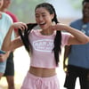 A young woman with braided hair smiles and holds her pigtails while wearing a pink Ron Jon Surf Shop crop top and matching shorts outdoors.