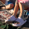 A person with sandy feet wearing light pink slide sandals sits on a blanket at the beach, with another person partially visible in the background.