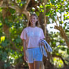 A woman in a pink Ron Jon t-shirt and checkered shorts walks outdoors, holding a gray sweatshirt, with green foliage in the background.