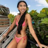 A woman in a red and white bikini stands near a fence and green foliage, looking to the side. Sunny day with a partly cloudy sky.