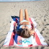 Woman in a blue checkered bikini lies on a red and white striped Ron Jon towel on a sandy beach, facing the sky.