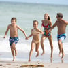 A boy, a girl, a woman, and a man in swimsuits run together along the shoreline at a beach on a sunny day.