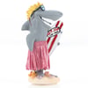 A cartoon shark figurine wears sunglasses and a pink grass skirt, holding a red and white striped surfboard standing on a sandy base with small sea decorations.