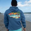 A person wearing a blue Ron Jon Surf Shop Cocoa Beach hoodie stands on a sandy beach, facing the ocean under a partly cloudy sky.