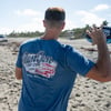 A person wearing a blue Ron Jon Surf Shop T-shirt stands on a sandy beach, holding a football and preparing to throw it. Trees and clouds are visible in the background.