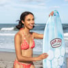 Woman in a red bikini smiles while standing with a blue surfboard at a beach. Waves are visible in the background.