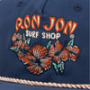 Close-up of blue cap featuring Ron Jon Surf Shop logo with orange and green flower embroidery, and a gold braided trim at the base.