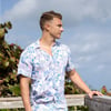 Man in a floral shirt leans on a wooden railing, looking to the side. Sky and foliage in the background.