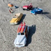 Toy cars and figures arranged on sand, including a yellow convertible, red and blue cars, and a white car with surfboards. Two miniature figures under an umbrella are nearby.