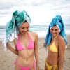 Two women in bikinis stand on a sandy beach with towels wrapped around their heads, smiling with the ocean and cloudy sky in the background.