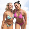 Two people smiling, wearing colorful bikinis; one in a floral pattern and the other in solid pink, standing against a backdrop of a cloudy sky.